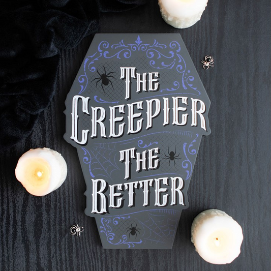 A coffin-shaped Halloween sign reading 'The Creepier The Better' in white gothic lettering with purple decorative flourishes and spider motifs, displayed on a dark wood surface with candles and small spiders around it.