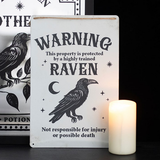 Decorative sign with a raven design and text, placed next to a candle on a dark surface.