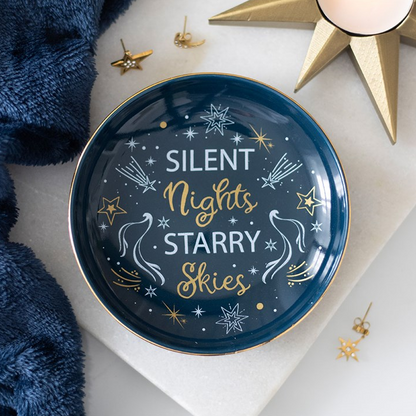 A round decorative tray with the phrase 'SILENT Nights STARRY SKIES' in white and gold lettering, styled on a white surface with gold star-shaped candles, small gold earrings, and a navy blue textured fabric.