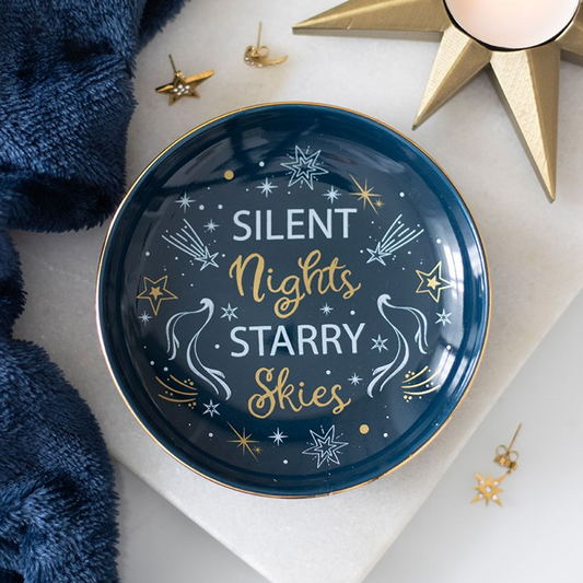 A round decorative tray with the phrase 'SILENT Nights STARRY SKIES' in white and gold lettering, styled on a white surface with gold star-shaped candles, small gold earrings, and a navy blue textured fabric.