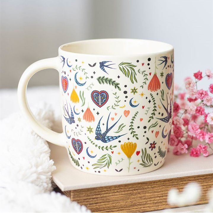 Decorative mug with floral and bird pattern on a white surface with flowers in the background
