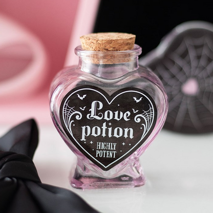 A small heart-shaped glass potion bottle with a cork stopper and a black heart-shaped label reading 'Love potion' and 'Highly Potent.' The bottle is photographed on a white surface with a pink and black themed background.