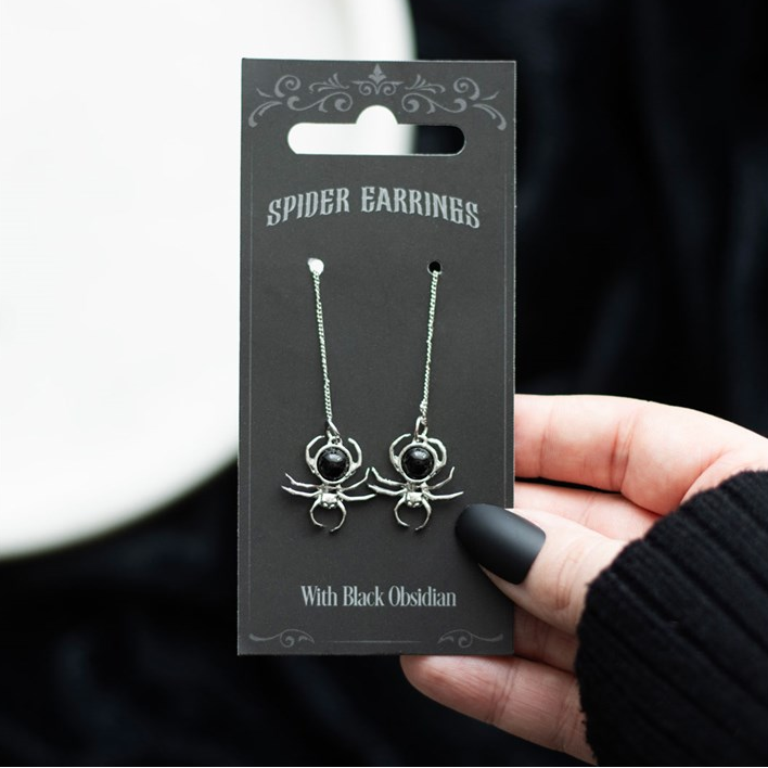 Spider earrings on a black card held by a hand with black nail polish.