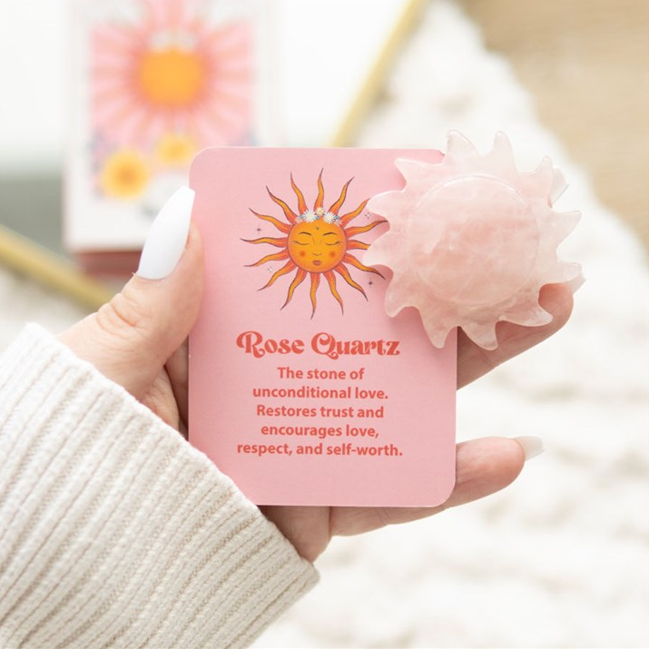 Hand holding a pink rose quartz stone with sun design and text.
