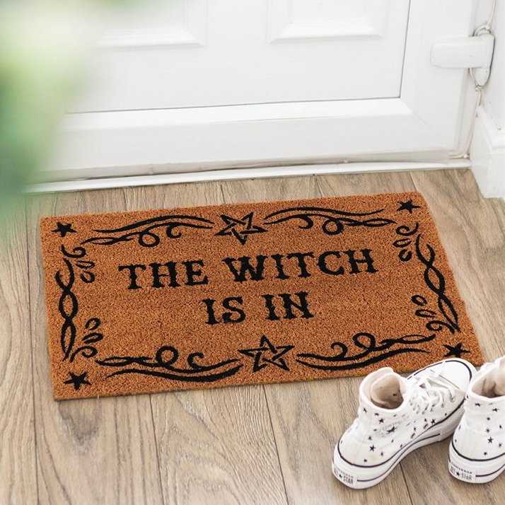 Doormat with 'The Witch is In' text on a wooden floor, with white shoes at the bottom.