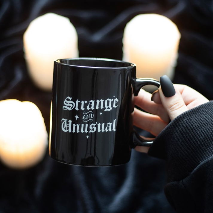 Black mug with 'Strange and Unusual' text held by a hand against a dark background with candles.