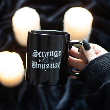 Black mug with 'Strange and Unusual' text held by a hand against a dark background with candles.