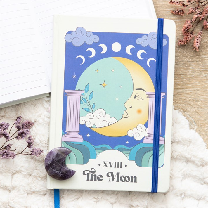 Notebook with 'The Moon' tarot card design on a white surface with flowers.
