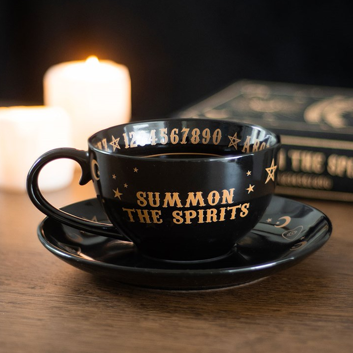 Black cup and saucer set with 'Summon the Spirits' text on a wooden surface with candles in the background.