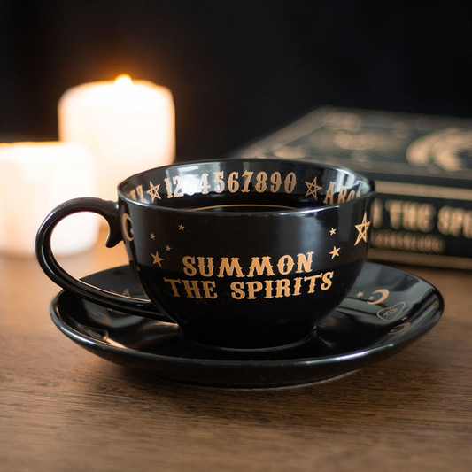 Black cup and saucer set with 'Summon the Spirits' text on a wooden surface with candles in the background.