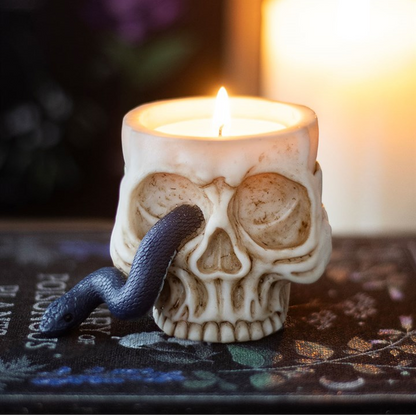 A skull-shaped candle holder filled with a lit white candle, with a dark blue snake draped around the left side, set on a dark patterned surface against a softly blurred warm background.