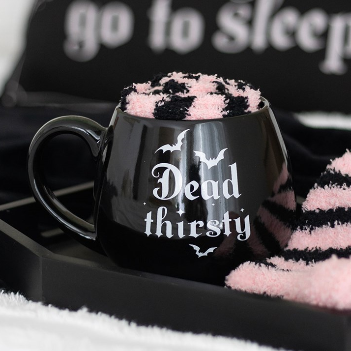 Black mug with 'Dead thirsty' text on a black surface with pink and white striped fabric.