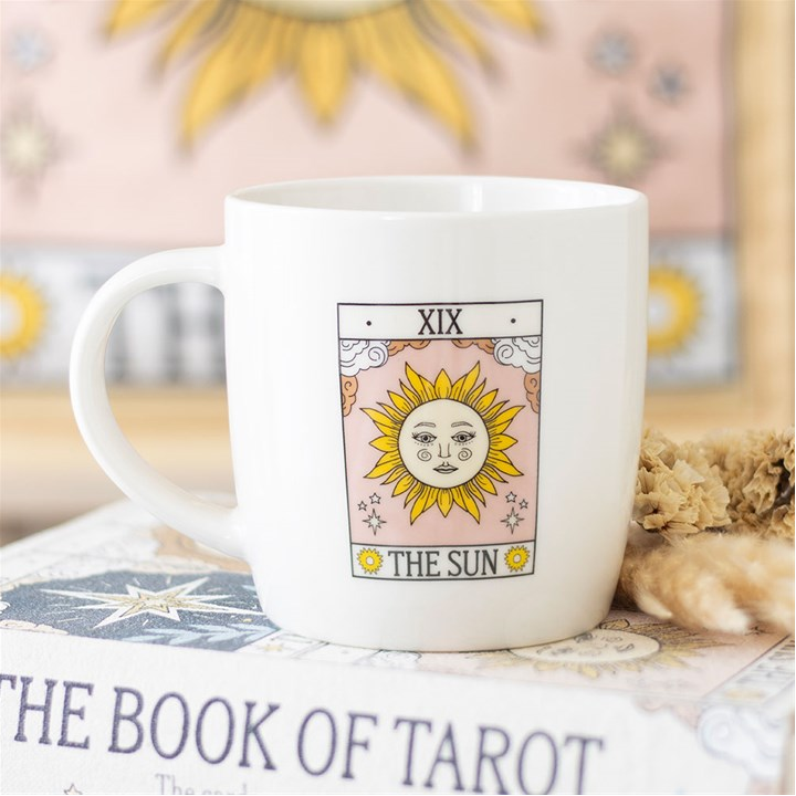 White mug with 'The Sun' tarot card design on a surface with tarot cards and flowers.