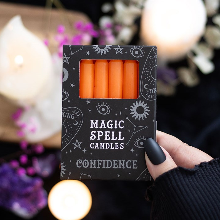 Pack of 'Magic Spell Candles' labeled 'Confidence' held by a hand with a blurred candle and flowers in the background.