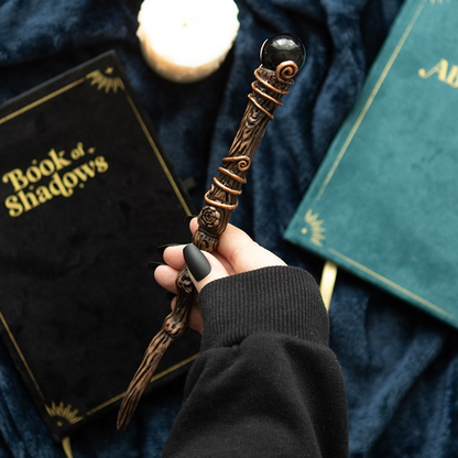 Hand holding a decorative wand over books titled 'Book of Shadows' and 'Albus'.