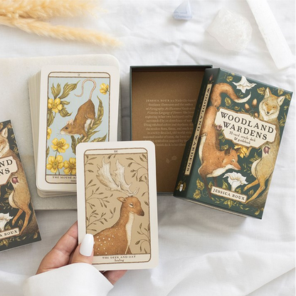 A styled flat-lay of a woodland-themed deck of cards and its box on white fabric, with a hand holding one card. The card and box illustrations feature deer, birds, and other forest animals in warm earthy tones.