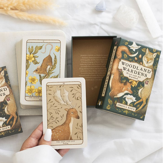 A styled flat-lay of a woodland-themed deck of cards and its box on white fabric, with a hand holding one card. The card and box illustrations feature deer, birds, and other forest animals in warm earthy tones.