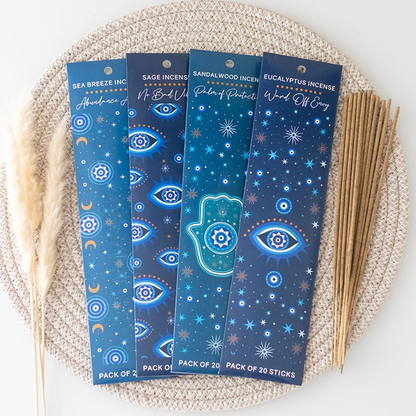 Four packaged incense sticks arranged on a woven beige mat with dried grass stems and a bundle of wooden sticks beside them. The packages are dark blue with celestial and Hamsa designs, and the incense sticks are light tan.