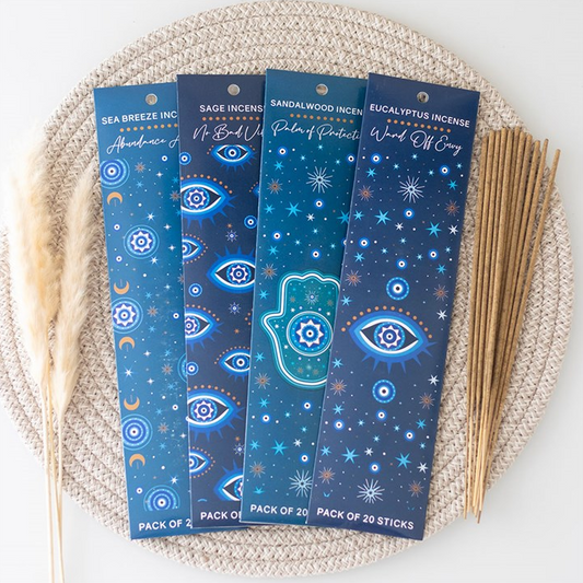Four packaged incense sticks arranged on a woven beige mat with dried grass stems and a bundle of wooden sticks beside them. The packages are dark blue with celestial and Hamsa designs, and the incense sticks are light tan.