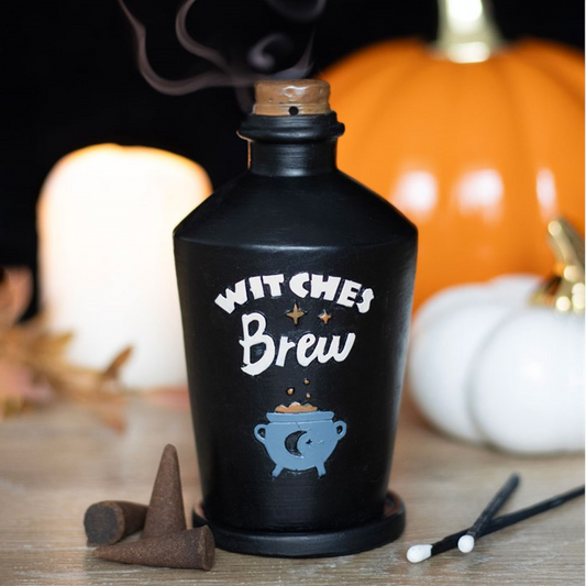 A black witch-themed bottle candle labeled 'WITCHES Brew' with a blue cauldron graphic, shown lit on a tabletop with pumpkins, a white pillar candle, and matchsticks in a cozy autumn setting.