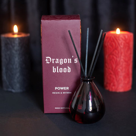 A reed diffuser bottle with black reeds stands in front of its burgundy box, with two lit candles blurred in the background on a dark surface.