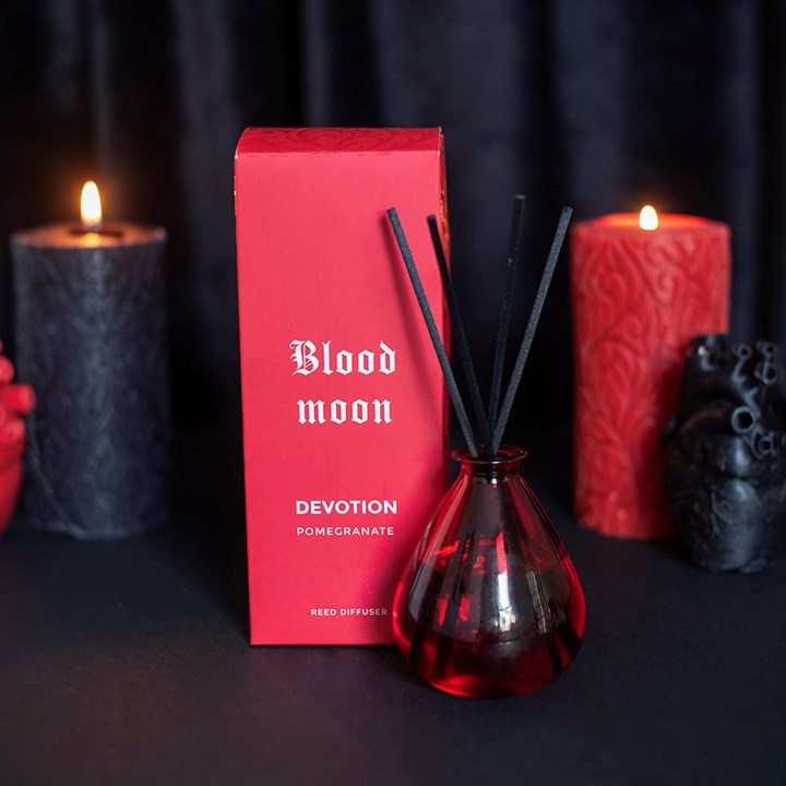 A reed diffuser set with a red glass bottle, black reeds, and a red box labeled 'Blood moon' and 'Devotion Pomegranate', styled with lit candles on a dark background.