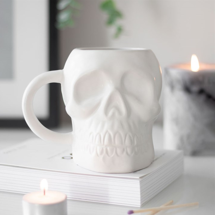 White skull-shaped mug on a surface with candles and books in the background