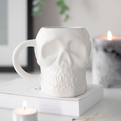 White skull-shaped mug on a surface with candles and books in the background