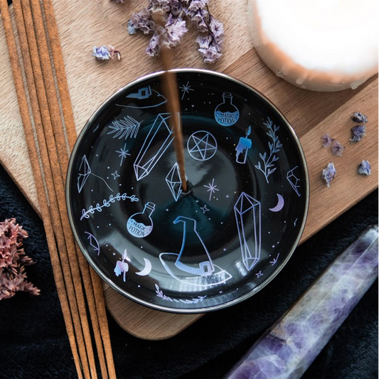 A dark ceramic bowl with white occult and mystical illustrations, including crystals, potions, and a pentagram, shown on a wooden board with dried flowers and a candle.