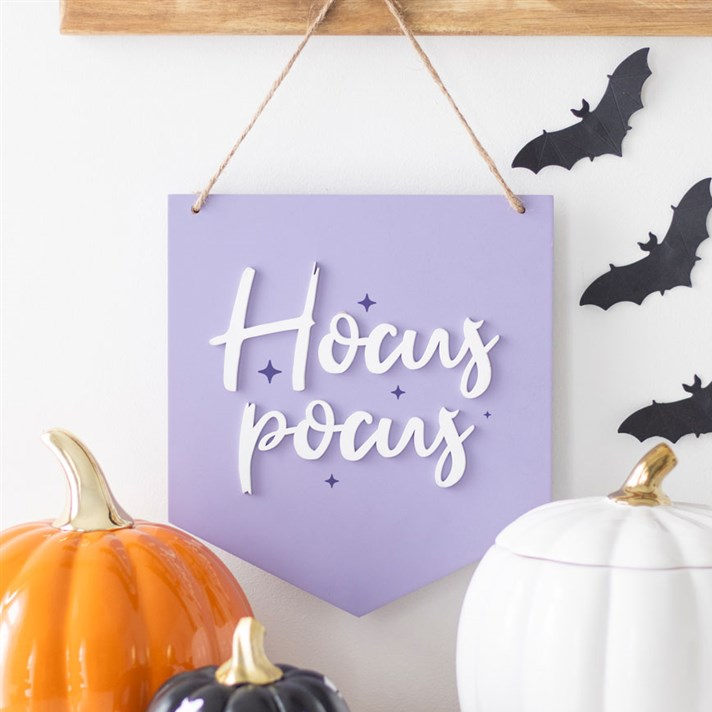 A lavender pennant-shaped hanging wall sign with the words 'Hocus Pocus' in white script, displayed on a wall with black bat cutouts, a wooden rod, and decorative pumpkins below.