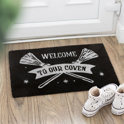 Black doormat with 'Welcome to our coven' text and broomsticks on a wooden floor.