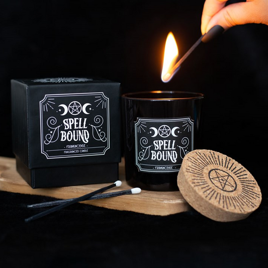 Black candle labeled 'Spell Bound' being lit with a match, surrounded by packaging and accessories on a dark background.