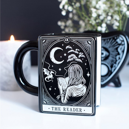 Black mug with 'The Reader' tarot card design featuring a silhouette of a person reading under a moonlit sky, with a blurred candle and plant in the background.