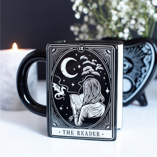 Black mug with 'The Reader' tarot card design featuring a silhouette of a person reading under a moonlit sky, with a blurred candle and plant in the background.